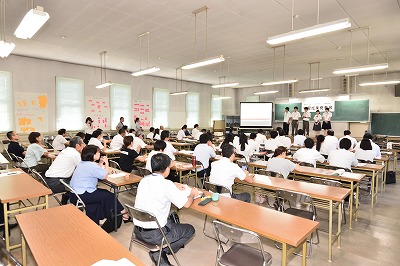 写真：グループごとの発表。ステージにたつ1グループ。発表を聞く他のグループや高校教員や市関係者