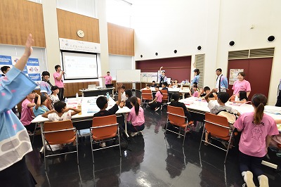 写真：お金についてのクイズを出す学生たち。子どもたちは手を挙げて答える