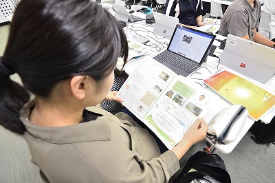 写真：できあがった情報紙を見て、片方の班の発表を聞く学生。後ろ姿のアップ