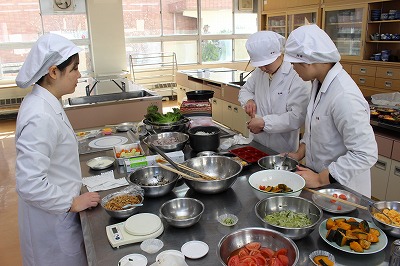 写真：試食を繰り返す学生3人。調理台に材料がいっぱい並ぶ
