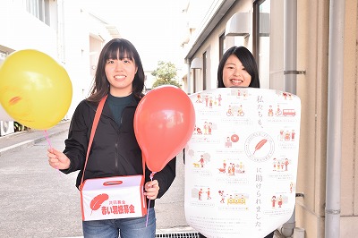 写真：募金箱と風船をもった学生1名とポスターを持った学生1名
