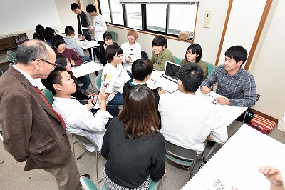 写真：パソコンを前に説明を受ける3年生、説明をする4年生、様子を見ている教員