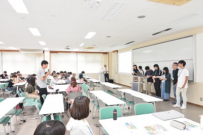 写真：アンケート調査の発表を終えた学生グループ1組が並んでいる。それを聞く学生と町内会の住民。一人の学生が立って質問をする姿