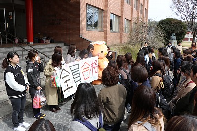 写真：全員合格と書かれた横断幕を持って、4年生の前に並んで立つ後輩学生とミマッパちゃん