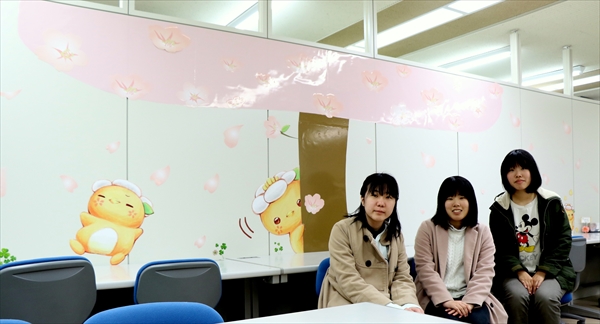 写真：写真：ミマッパホールの入り口の壁面にミマッパのシールが貼ってある様子。その前に座る3名の学生