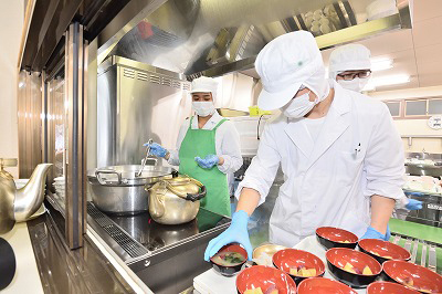 写真：汁物のさつまいもの味噌汁を器につぐ学生。量を確認する学生