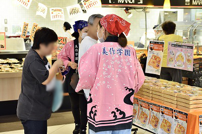 写真：試食を渡す学生。試食を受け取る家族連れ。こども2人とおとな2人