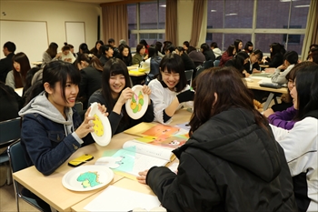 写真：紙皿で作ったおもちゃを持って遊び方を披露する3名の学生
