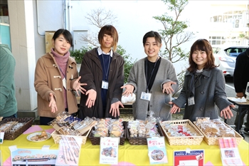 商品：売り場に並べた商品を前にポーズをとる4名の学生