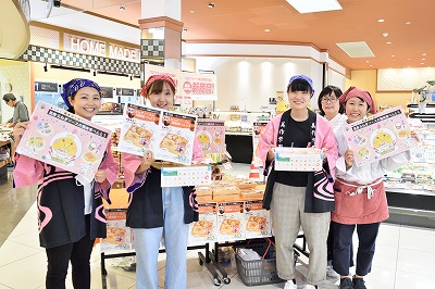 写真：マルイノースランド店の店内でマルイ担当者と学生3人、人見先生で販売ブースを前に記念撮影