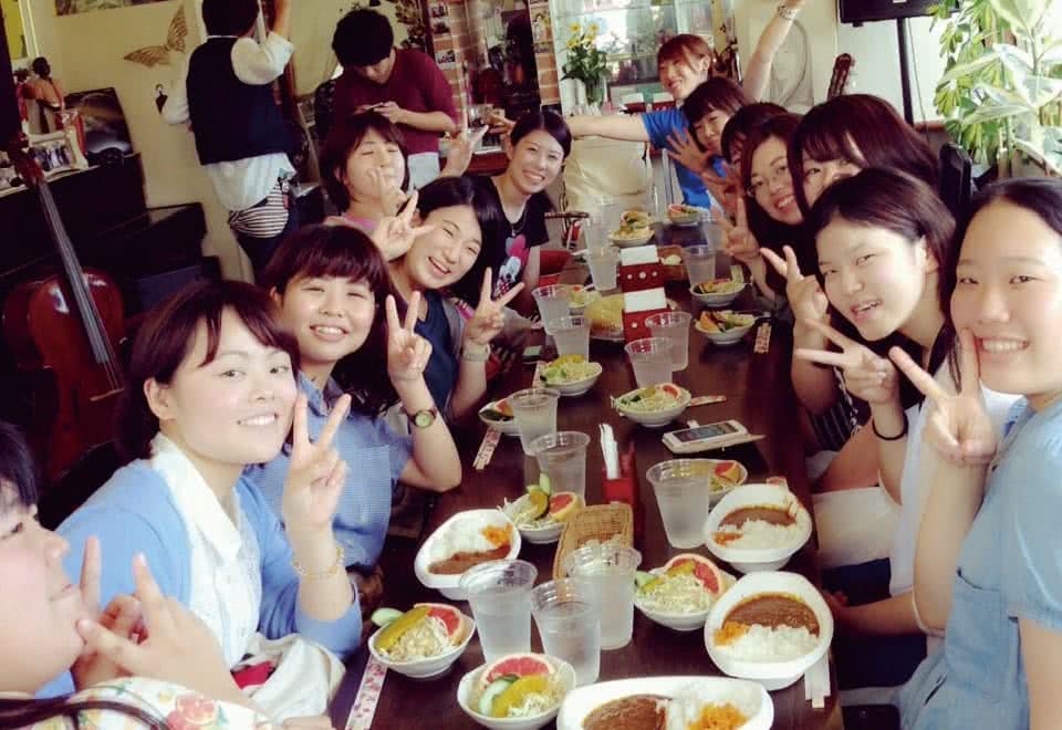 写真：懇親会の様子。テーブルの上にサラダとカレーが並んで、みんな笑顔でピースしています。