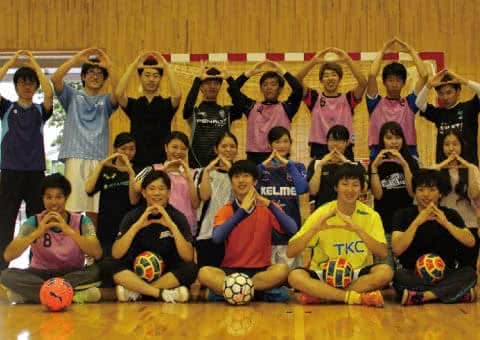 写真：体育館で両手を丸く合わせたポーズを取り写真に収まるフットサル同好会のメンバーたち