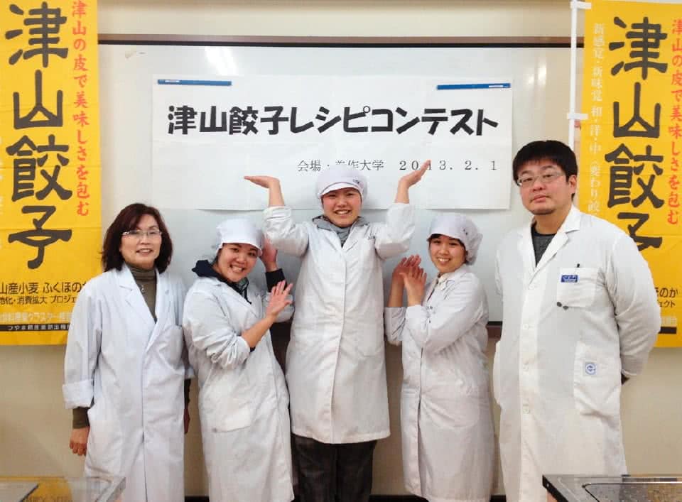 写真：「津山餃子」と書かれた旗を左右に、中央に「津山餃子レシピコンテスト」と書かれた紙が貼られたホワイトボードの前で白衣を着て並ぶつやま夢みのりグループの5人