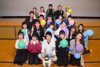 写真：体育館の壇上の前に集合し、両手を広げたポーズで集合写真に収まる学友会のメンバーたち