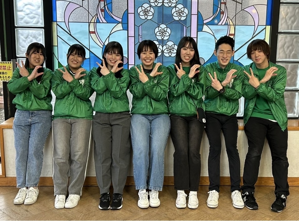 写真：ステンドグラスを背景に、緑色のジャンパー姿でポーズをとる7人の学生