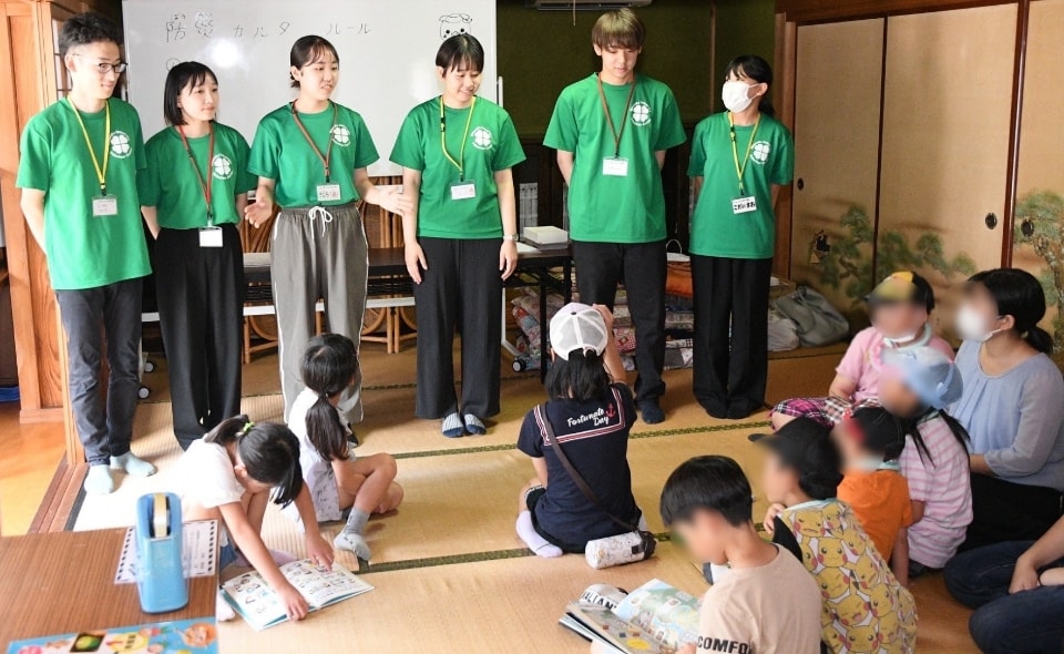 写真：和室で、緑のTシャツを着た学生たちが並んで立ち、座っている子どもたちに説明している様子