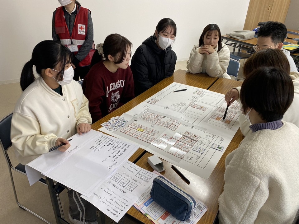 写真：机いっぱいに広げた図面を囲み、真剣な表情で議論する学生たち。後ろには赤十字のスタッフが立って見守っている