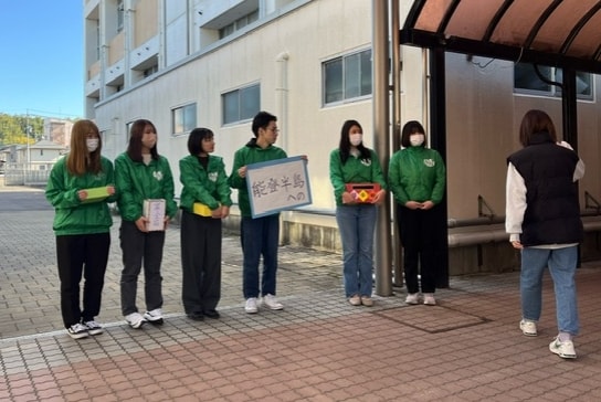 写真：能登半島への募金を呼びかける緑のジャンパーを着た学生グループと、その前を通る学生。校舎前の屋外で撮影