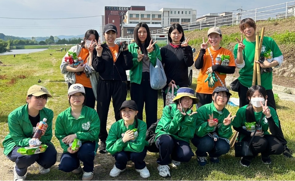 写真：河川敷でごみ拾いを終え、緑のジャンパーやオレンジ色のTシャツ姿で笑顔とピースサインを見せる学生たち