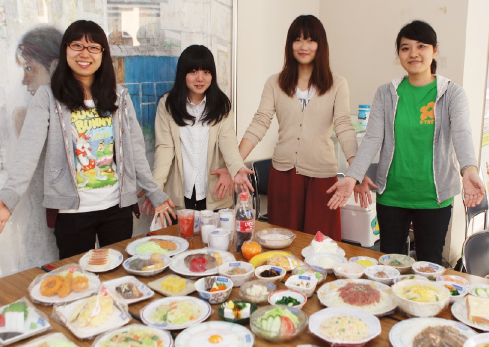 写真：テーブルに並べれれたたくさんの料理を両手を広げたポーズで紹介する4人の女子食育サークルメンバー