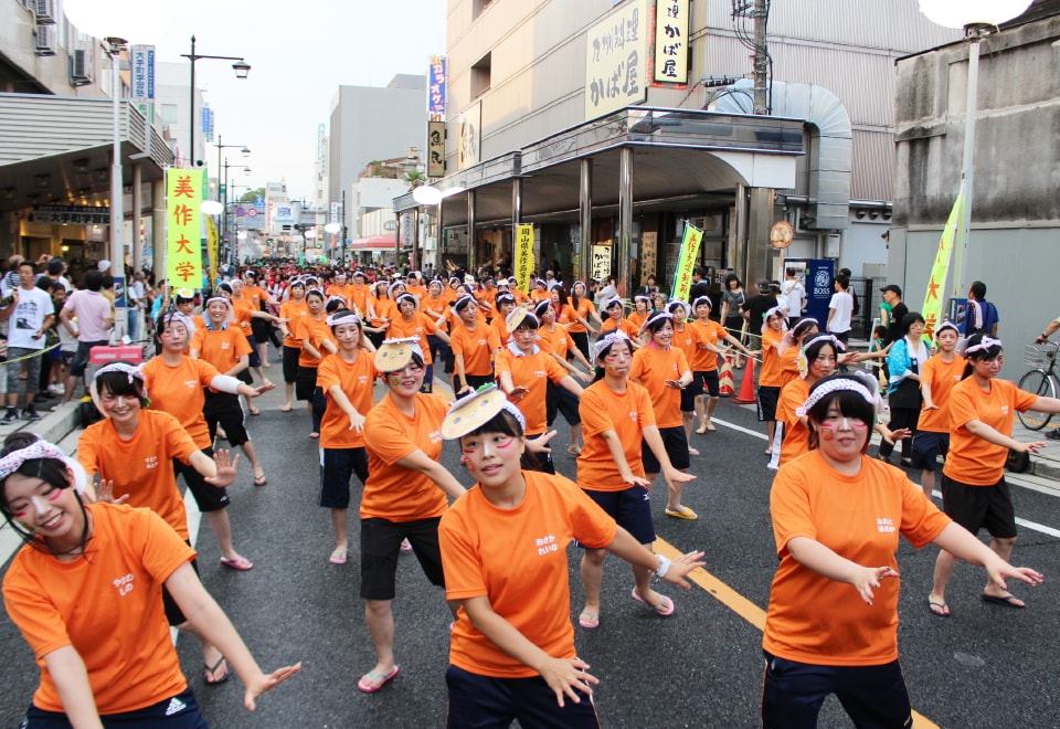 写真：オレンジ色のTシャツを着て、頭にミマッパちゃんのお面を付け、顔にメイクをして一生懸命ごんごおどりを踊る幼児教育学科の学生たち