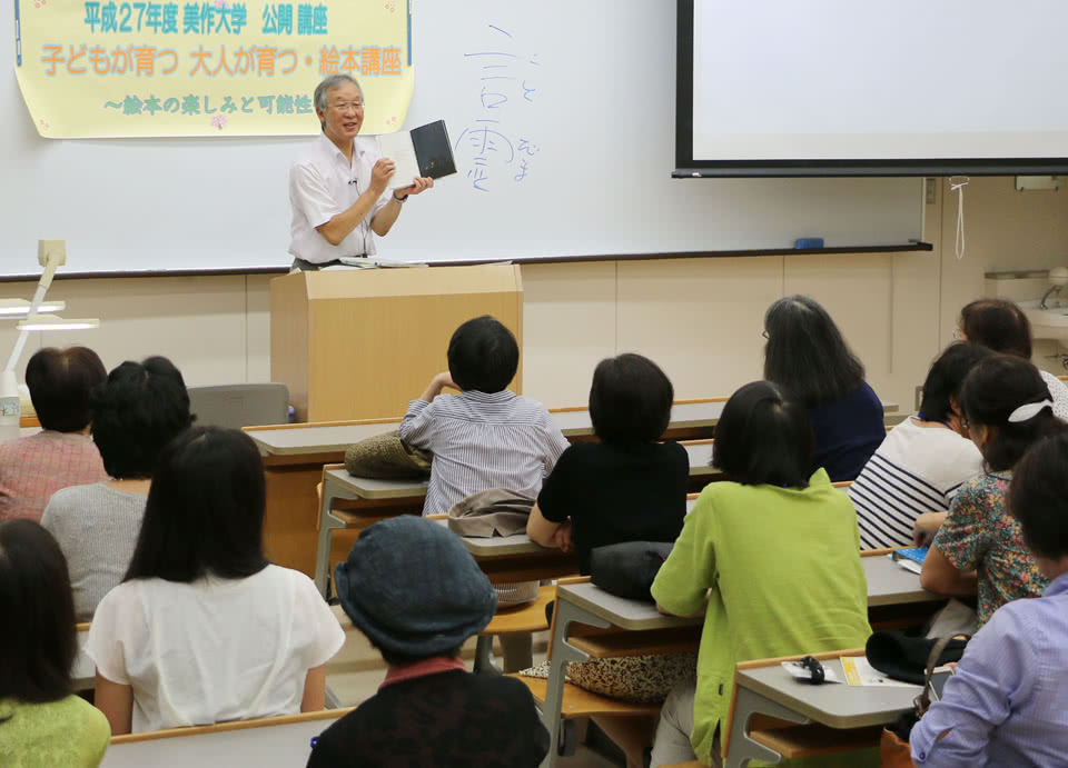 写真：「平成27年度 美作大学 公開講座 子どもが育つ 大人が育つ 絵本講座 ～絵本の楽しみと可能性～」と講座名が書かれた紙が貼られたホワイトボードの前で公開講座を行う講師と、受講する地域の人々。