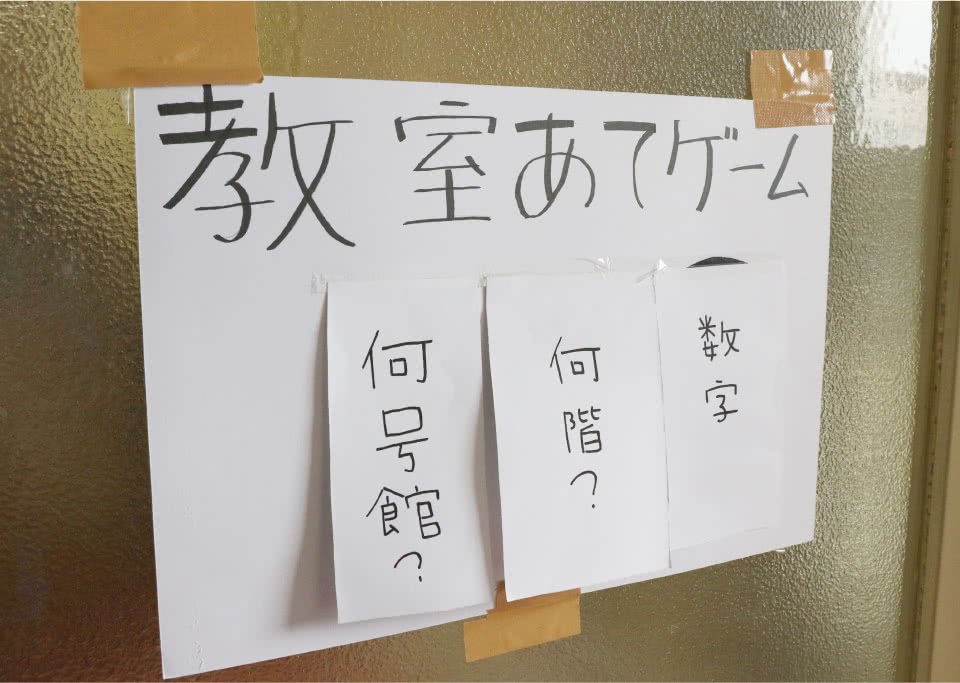 写真：教室あてゲームと書いた画用紙に「何号館？」「何回？」「数字」をと書いた紙が貼られたものをガムテープでガラス戸教室に貼ってあるもの。