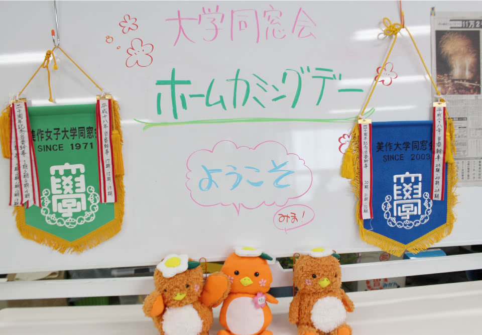写真：ホワイトボードに書かれた「大学同窓会 ホームカミングデー ようこそみま！」という文字と、美作大学同窓会の旗2つと3体のみまっぱちゃんのぬいぐるみ