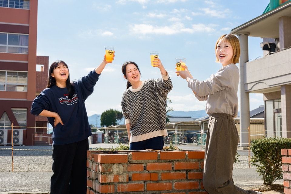 レンガの囲いの前で楽しそうに乾杯する女性3人。快晴の空と学校の校舎が背景に見える
