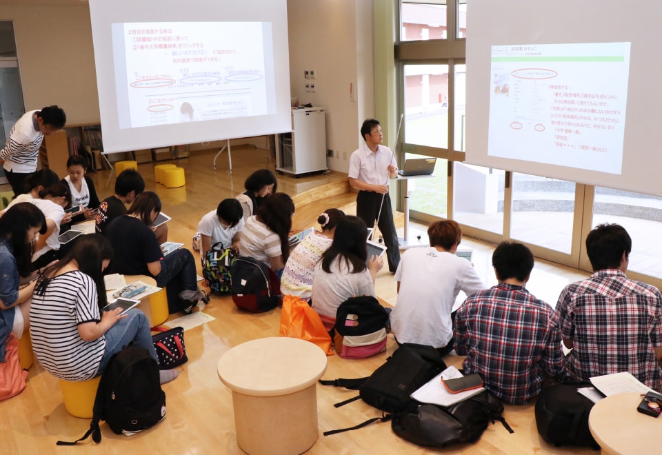 写真：サークルコモンズのステージ中央でプロジェクターを使った授業を行っている教員と、それを聞いている学生たち。
