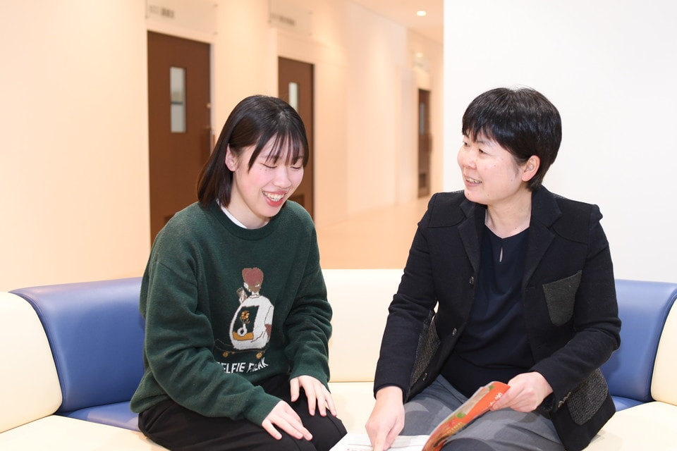 写真：明るい廊下のソファで、学生と職員が資料を手に楽しそうに会話している様子