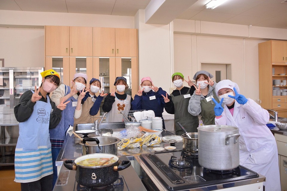 写真：調理実習室でエプロンや帽子、マスクをつけてピースサインをする学生グループ。前には調理中の鍋や食器が並んでいる